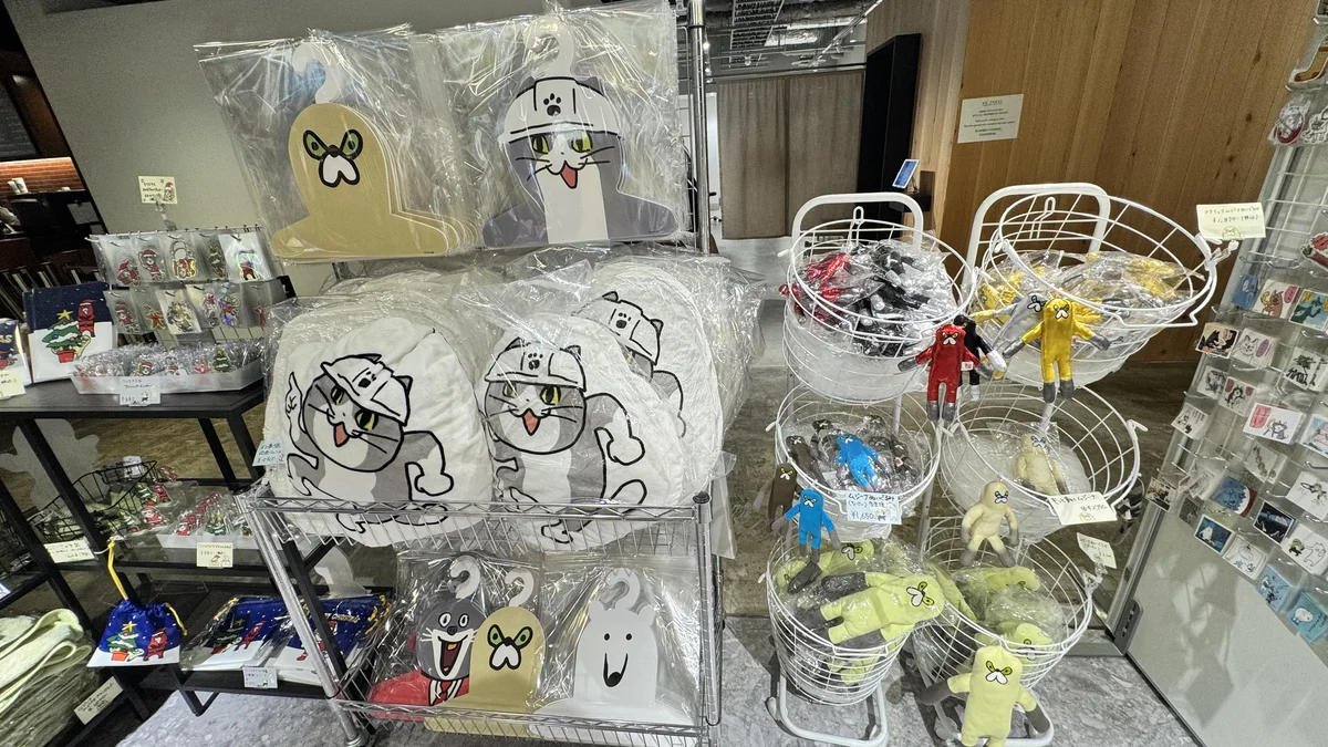 モナジアナの仕事猫クッションとムジーナのぬいぐるみが並ぶ棚。カラフルなぬいぐるみがワイヤーバスケットに詰まっている